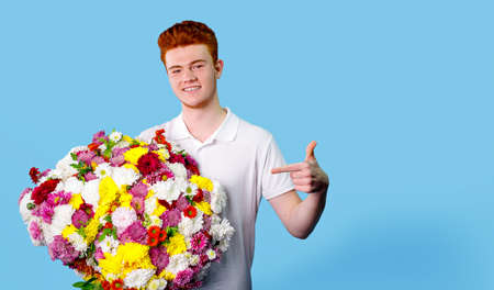 Happy young man holding a huge beautiful bouquet of flowers and pointing at it with his indicate finger over blue background.の写真素材