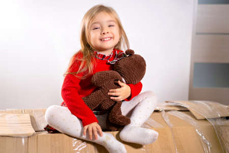 Smiling pretty girl blonde sitting on the cardboard big box tied with scotch tape holding in her hands favorite soft toy bear.の写真素材