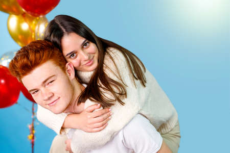 Photo of a young brunette woman climbed up on the back of her red hair man embracing him on the neck making fun over blue background and red gold balloons.の写真素材