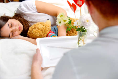 A man holding the tray with a vase of flowers and red heart shape from cardboard to make romantic breakfast for his girlfriend sleeping hugging a bear toy.の写真素材