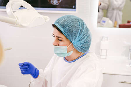 Side view photo of a woman dentist in uniform, medical mask and gloves ready begin to work with a patient in stomatology clinic.の写真素材