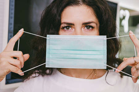 Close up photo of a brunette woman looking at the camera covering her face with a medical mask holding in her hands.の写真素材