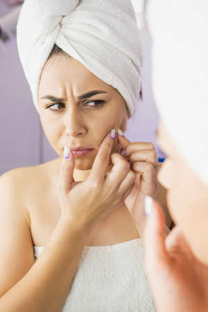Dissatisfied stressed depressed young woman with a towel on her head looking in the mirror touch cheek worried about skin wrinkle, cosmetology facial treatment.の写真素材