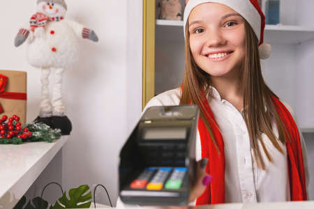 Happy young woman in Santa hat and red scarf holding payment terminal with office and holidays concept.の写真素材