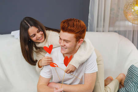Pretty young lady in warm sweater sitting on her knees on the sofa behind her boyfriend and flirting showing him heart shape flowers.の写真素材