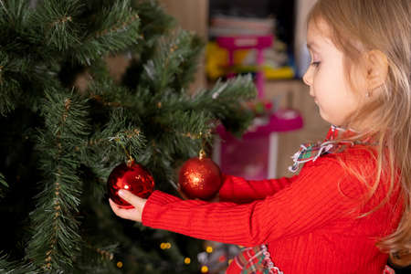 Little pretty girl blonde in red standing near the Christmas tree and touching two red decorative bubbles on the tree looking at them.の写真素材
