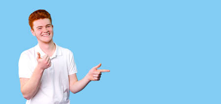 A red hair color boy smiling wearing white t-shirt gesturing with his hands indicates with his fingers to the back space on the blue background.の写真素材