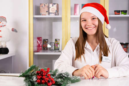 Cheerful business young woman wearing Santa hat standing at her workplace office manager with a brunch of tree on the table.の写真素材