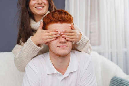 Young man sitting on the sofa and his girlfriend standing behind him closed his eyes with her palms smiling.の写真素材