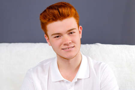 Close up photo of a young red hair man in white t-shirt sitting on a sofa over violet background posing for a camera.の写真素材