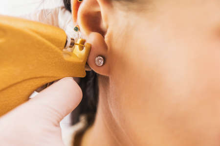 A doctor in sterile white medical gloves pierces the ears of a young girl in the medical office with a piercing gun.の写真素材