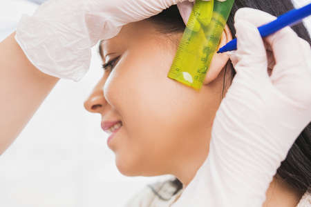 Cosmetologist hands holding ruler and pen to measure the place of a patient woman ear to pierce the earing.の写真素材