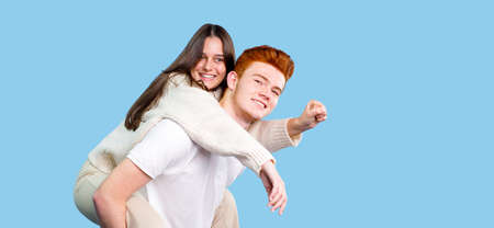 Two people brunette young woman and red hair young man making fun holding his girlfriend on the back over blue background, copy space.の写真素材