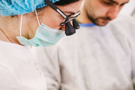 Stomatologist female doctor wearing dental loupe binoculars, medical mask and cap working together with an assistant man.の写真素材