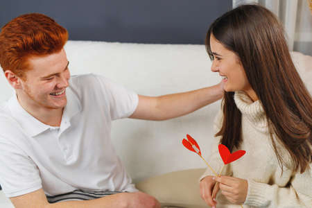 Loving couple sitting on the sofa opposite each other smiling, girl holding red heart cardboard flower and boy put his left hand on her shoulder.の写真素材