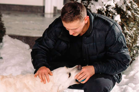 A man in winter clothes outside taking a blue little ball from the dog mouth to continue playing with the dog.の写真素材
