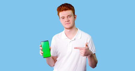Photo of cheerful and attractive guy in white t-shirt, having smart phone with green screen in hand,...
