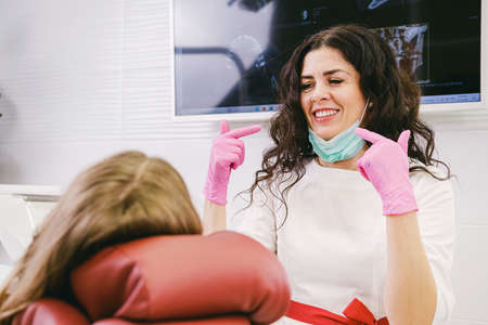 Close up photo of a dentist woman in white uniform with medical mask on her chin and pink gloves pointing with her two index fingers at her mouth.の写真素材