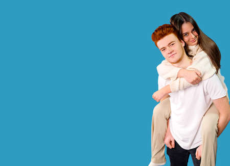Beautiful brunette girl climbed up on the back of her boyfriend and embracing him, he holding his girlfriend legs over blue background, copy space.の写真素材