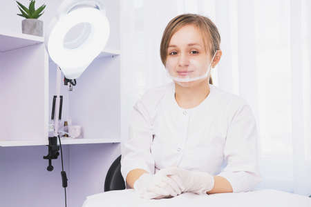 Young beautiful cosmetologist in a protective plastic mask facial screen and gloves sitting at a table in her cabinet in a beauty clinic studio waiting for client.の写真素材