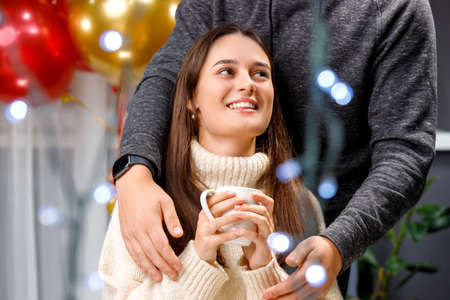Couple in love posing for a camera, young woman holding a cup in her hands and smiling and her boyfriend embracing her in the back with his hands.の写真素材