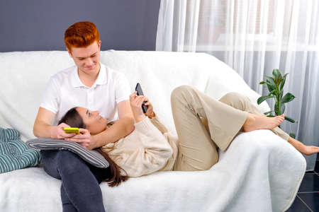 Young woman brunette lying on her boyfriend knees on the pillow on the sofa and both holding in their hands phones.の写真素材