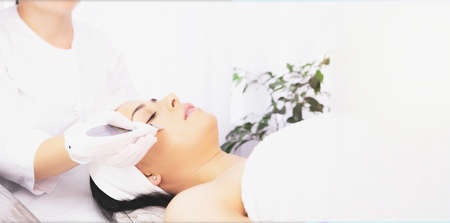Banner- long format. Female specialist doing facial for woman undergoes dermatologic procedures with ultra sonic scraber lying on a massage table in a clinic beauty salon.の写真素材