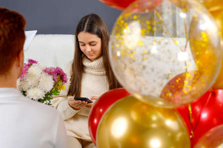 Young man making surprise for his girlfriend holding flowers in his hands and colored balloons while she sitting on a sofa looking at the phone.の写真素材
