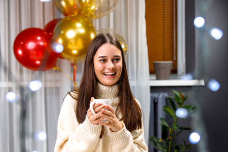 Happy young woman brunette in warm white sweater holding a cup with two hands having balloons over background.の写真素材