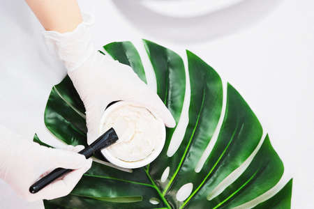 Close up photo of a face mask cream in a jar on a green big monstera leaf hold by a cosmetologist in a beauty clinic salon.の写真素材