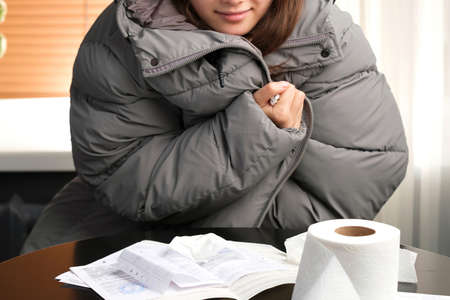 Cropped photo of a young girl in winter coat sitting at the table with payments invoices and paper on it, turning-off heating.の写真素材