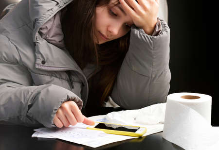 Upset young frozen woman puts on a hood sits at the table in cold room during turning off home heating in winter season and looking at the invoices and smart phone.の写真素材
