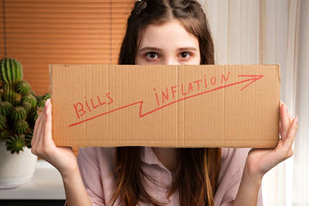 Young woman holding with her two hands cardboard with an inscription and arrow up covering her face, payment problem concept.の写真素材