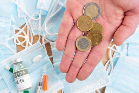 Cropped photo of a hand with coins and many medical masks with vial of vaccine and sterile syringe on the table.の写真素材