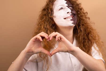 A beautiful girl with red hair and blue eyes painted with the Qatar flag on her face shows a heart with her hands. Isolated on a brown backgroundの写真素材