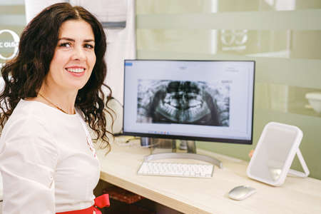 Side view photo of a stomatologist woman sitting at the table with teeth snapshot on the computer in the clinic office.の写真素材