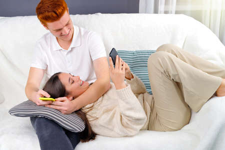 Loving couple spending time together holding smart phones in their hands and young pretty woman lies on the pillow on her boyfriend knees.の写真素材