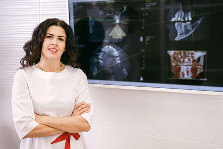Cheerful dentist woman in white uniform with cross hangs standing and smiling near the teeth snapshot on the computer.の写真素材