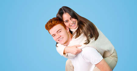 Two young people in love is having fun, pretty joyful lovely smiling girl is sitting on her boyfriends back isolated on blue background.の写真素材