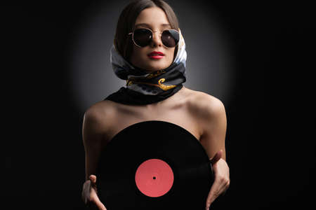 Fashionable pretty lady holding a vinyl record in her hands covering her chest wearing sunglasses and shawl on her head.の写真素材
