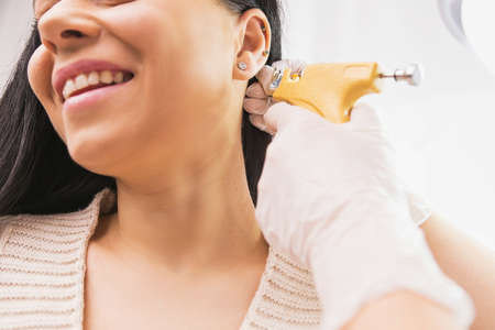 Cropped photo of a smiling woman patient getting ear piercing procedure by a specialist in medical gloves in the cosmetologist cabinet.の写真素材