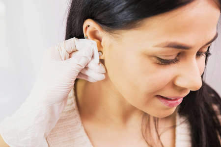 Side view photo of a beautiful brunette woman preparing to a piercing procedure and doctor in sterile gloves rubbing the ear with alcohol.の写真素材