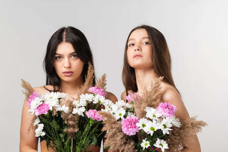 Close up photo of two beautiful girlfriends blonde and brunette with naked shoulders covering their breast with colorful bouquet of flowers.の写真素材