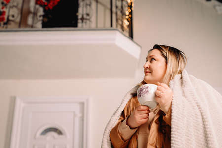 Side view photo of a woman outside in winter vacations looking up aside standing near the decorated house with garlands keeping a mug of hot drink.の写真素材