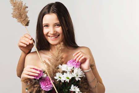 Close up photo of a happy young pretty lady covering her breast with flowers and somebody standing behind her with a brunch of reed.の写真素材