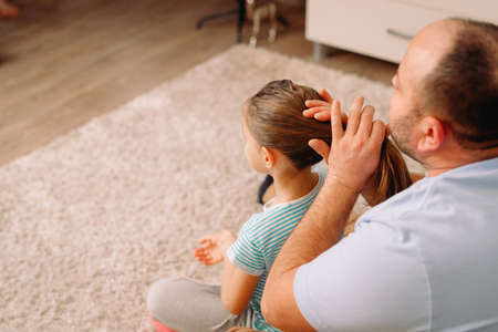 A caring father in a white t-shirt combs his little daughters hair while sitting on the sofa at home.の写真素材