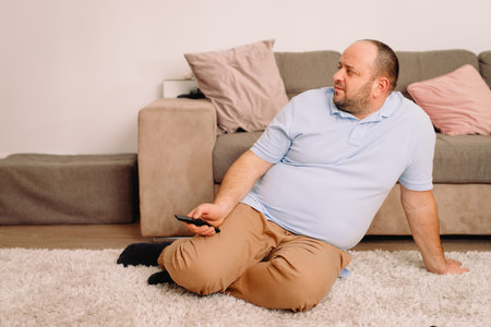 Father sitting on the carpet holding in his hands TV remote and pushing buttons and looking aside to...