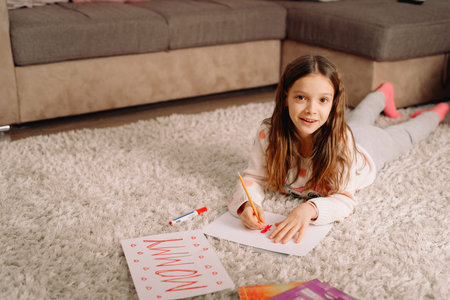 Pretty nice blond girl lying on the carpet at home writing on the paper word mommy using the red...