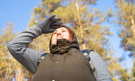 Young woman redhair in warm clothes an winter gloves keeping her hand on the forehead and looking into the distance outdoors.の写真素材