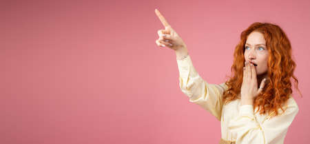 Amazing young girl with curly hairstyle redhead covering her mouth with a palm and pointing up with a finger of another hand on copy space over pink background.の写真素材